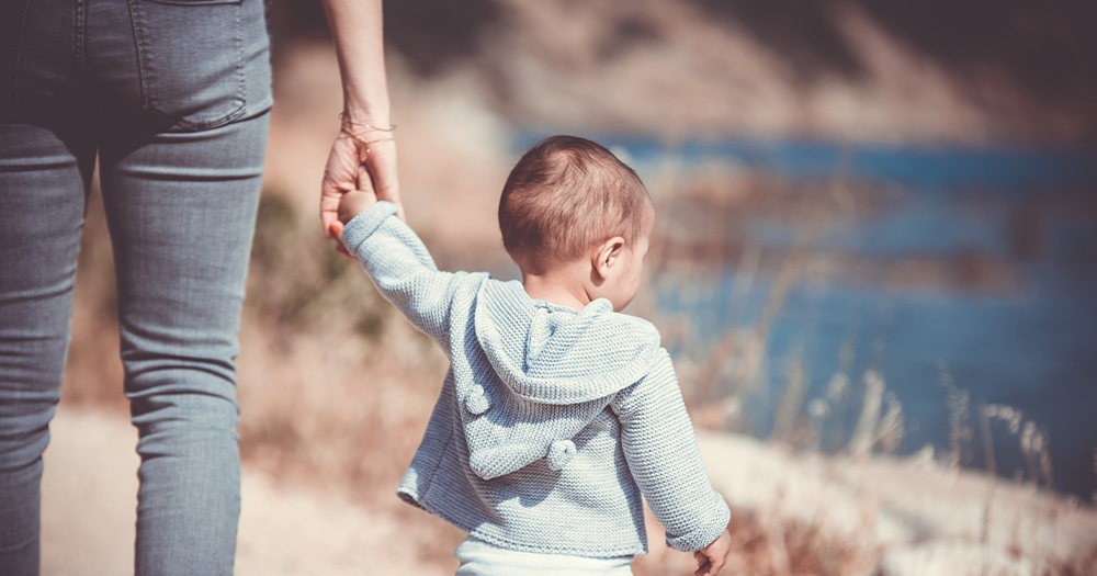 The torso of a woman holding a toddler's hand as they walk along a hill overlooking the sea