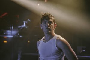 A young man in a vest poses moodily while lit by neon light