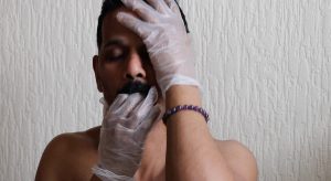 A topless man putting his rubber gloved hands in his mouth