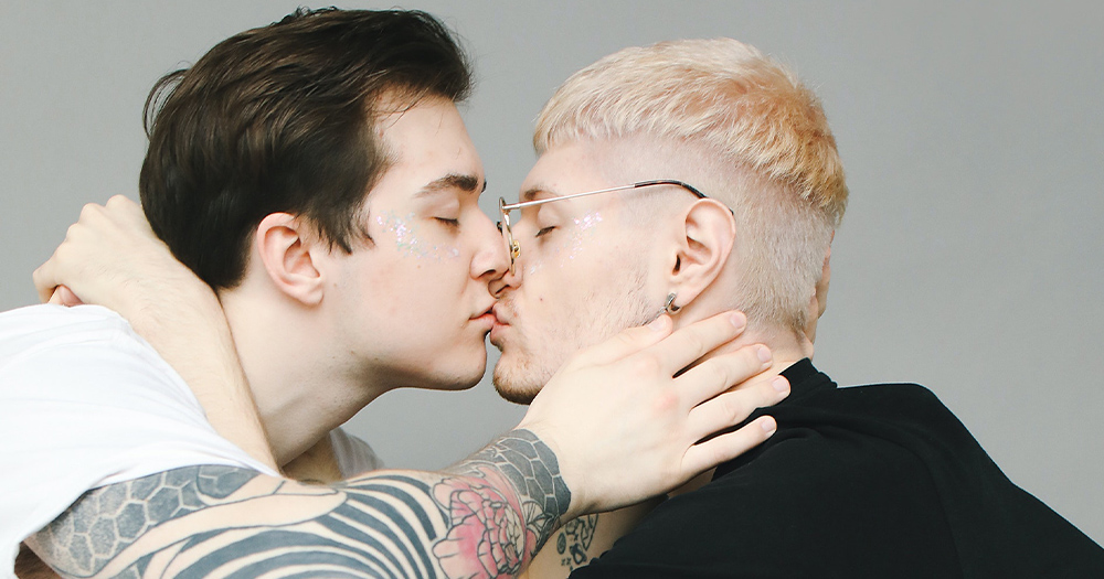 A heavily tattooed young man kisses another young men with bleached hair and glasses