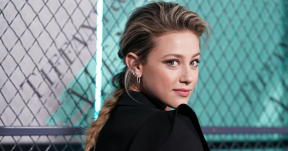 Actress Lili Reinhart, a young woman with long blonde hair in a plait, looks over her shoulder and smiles at the camera