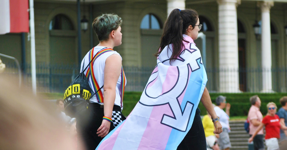 Two young people walking, one wrapped in trans flag