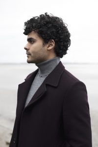 A curly haired Brazilian man in a coat stands on a grey beach