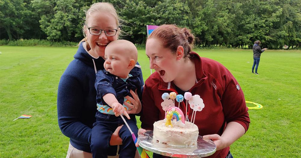 Two women hold a baby and a birthday cake while standing in a park