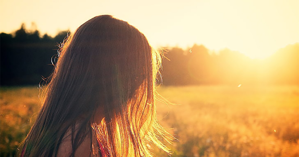 A woman with long hair covering her face in a field at sunset