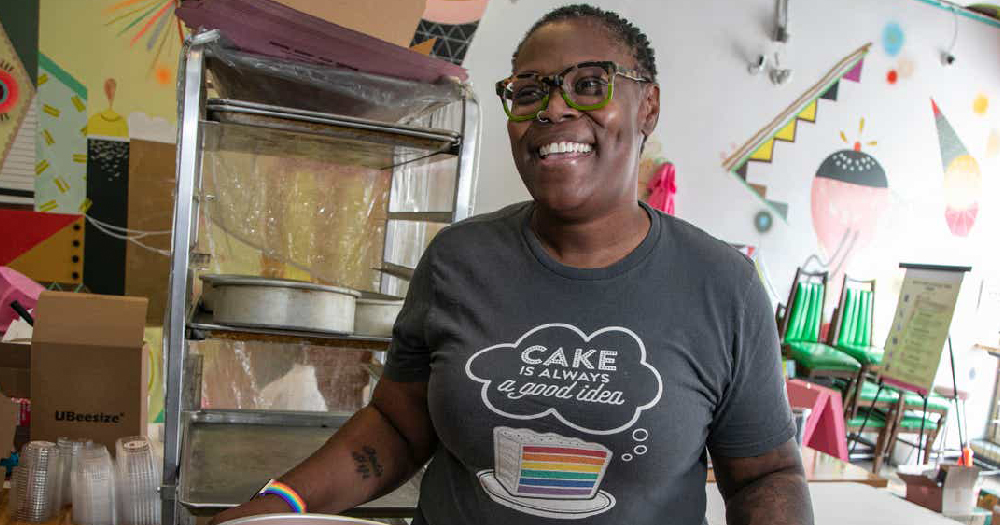 Queer baker, Anderson, smiling and wearing a shirt with a rainbow cake