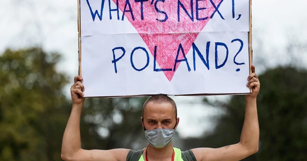 Poland LGBTQ+ man wearing a face mask holds a poster which has a pink triangle with text 'What's next Poland?'