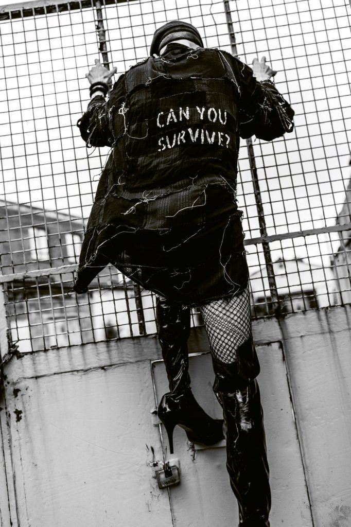 Drag queen Veda from behind holding during a lockdown photoshoot. She is wearing a jacket that reads "Can you survive"?