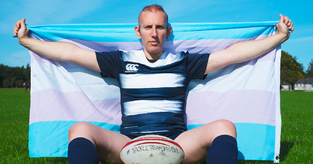 World Rugby. A trans woman sits on a rugby pitch wearing a rugby jersey and holding a trans flag behind her