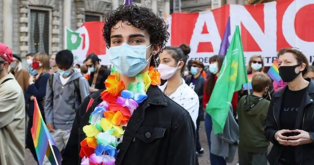 hate crime legislation Italy a masculine person with rainbow garland necklace stands in front of protesters wearing a face mask