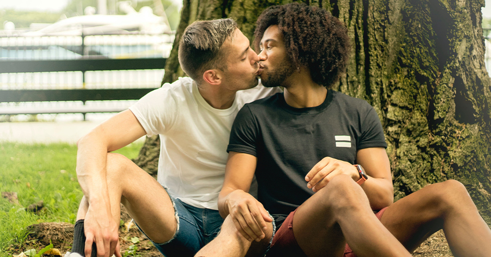 Two men kiss while sitting under a tree