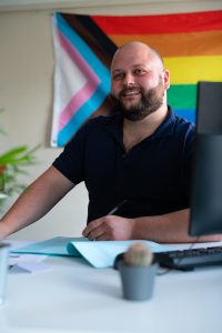 A smiling man sitting at a desk in front of a rainbow flag