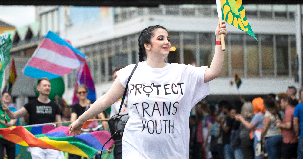 LGB Alliance Ireland debunked. A feminine person wears a t-shirt with 'Protect Trans Youth' written on it during the 2019 Dublin Pride Parade. Trans flags can be seen in the background.