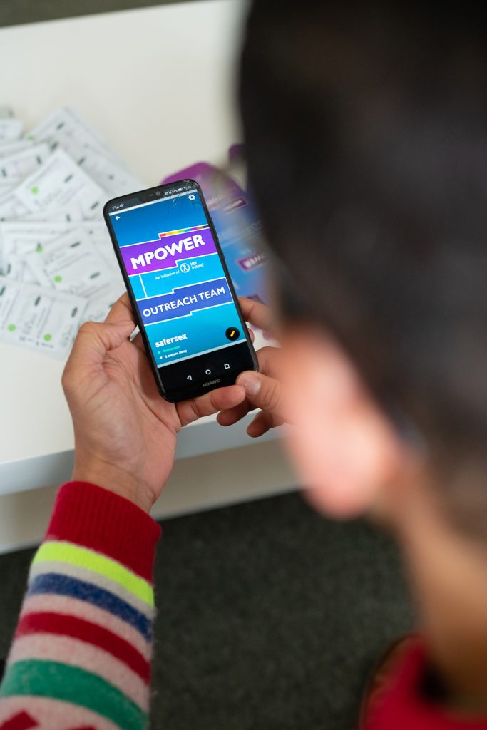 A man from behind holding a phone. The phone screen shows the MPOWER logo. Diego Caixeta from MPOWER talks about addressing the shame around sex.