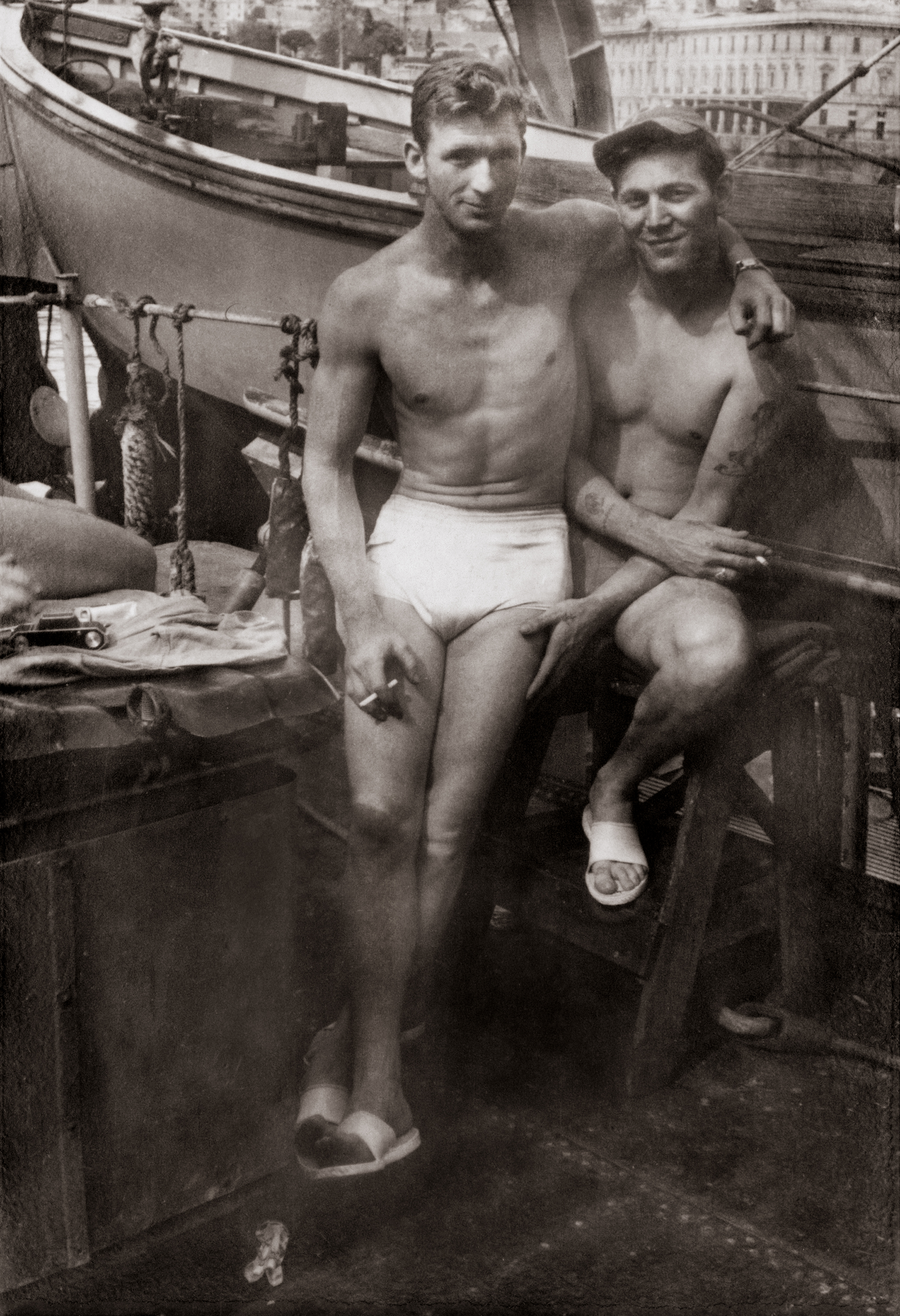 Two men in swim trunks leaning up against a boat, Loving, a history of men in love through photographs