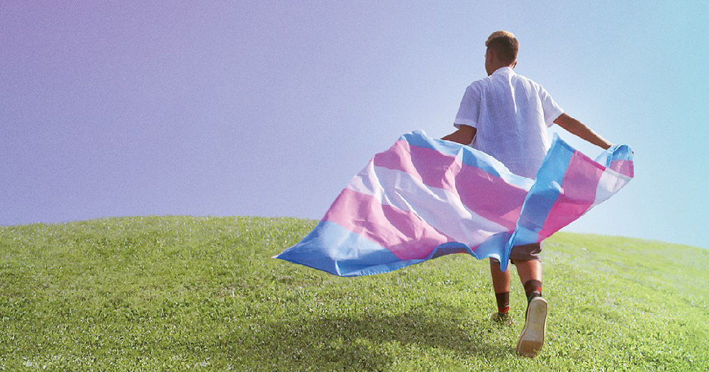 A young man runs through a field carrying a trans flag