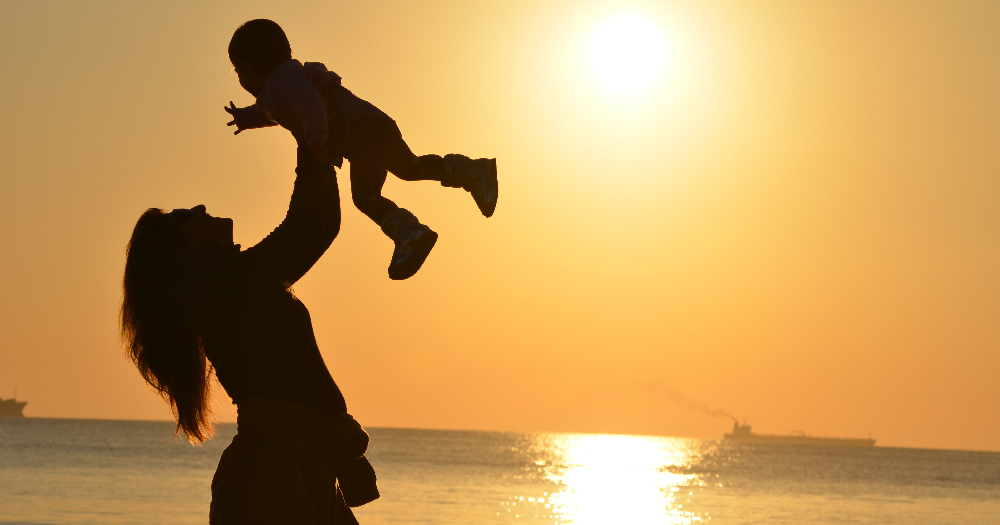 publicly funded IVF feminine person holding a baby in the air is sillouetted against a sunset on a beach