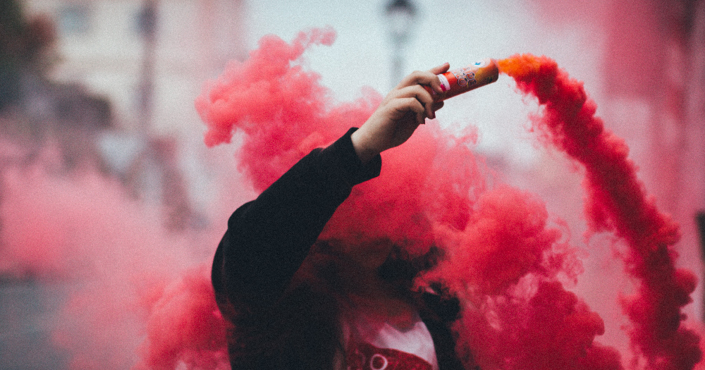 Far Right a person holds a smoke bomb which is billowing red smoke as they walk down a street