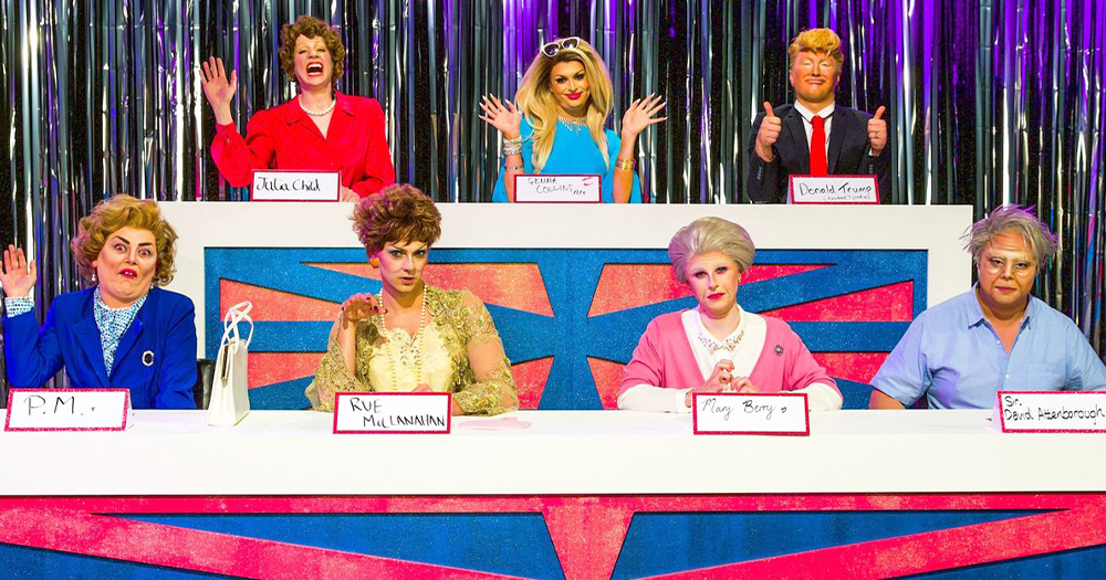 Seven people in fancy dress on a game show waving at the camera