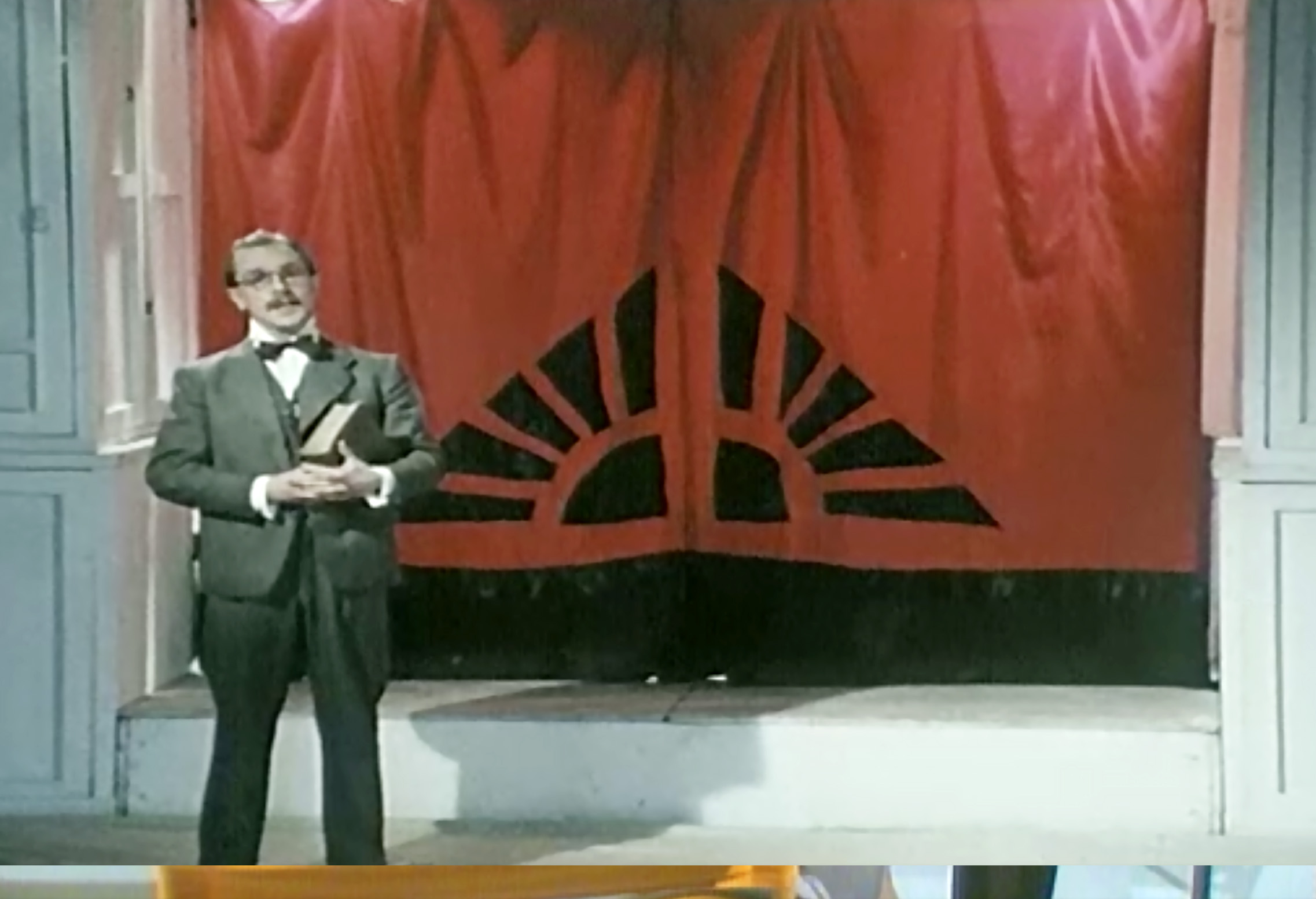 A man stands on a stage with a large book in his arms and a red curtain behind him.