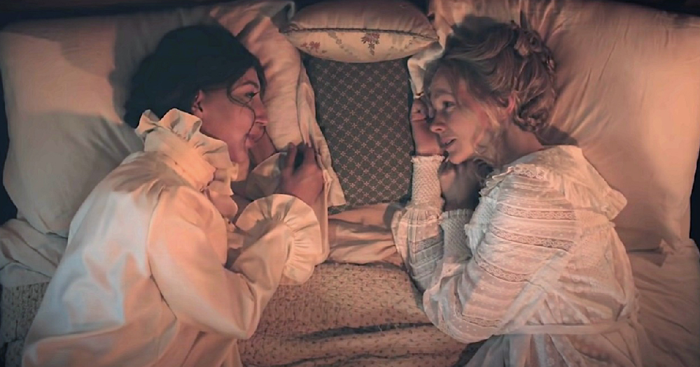 a Still image from Lesbian Period drama on SNL with two women lying in bed opposite each other in old fashioned nightdresses in candlelight