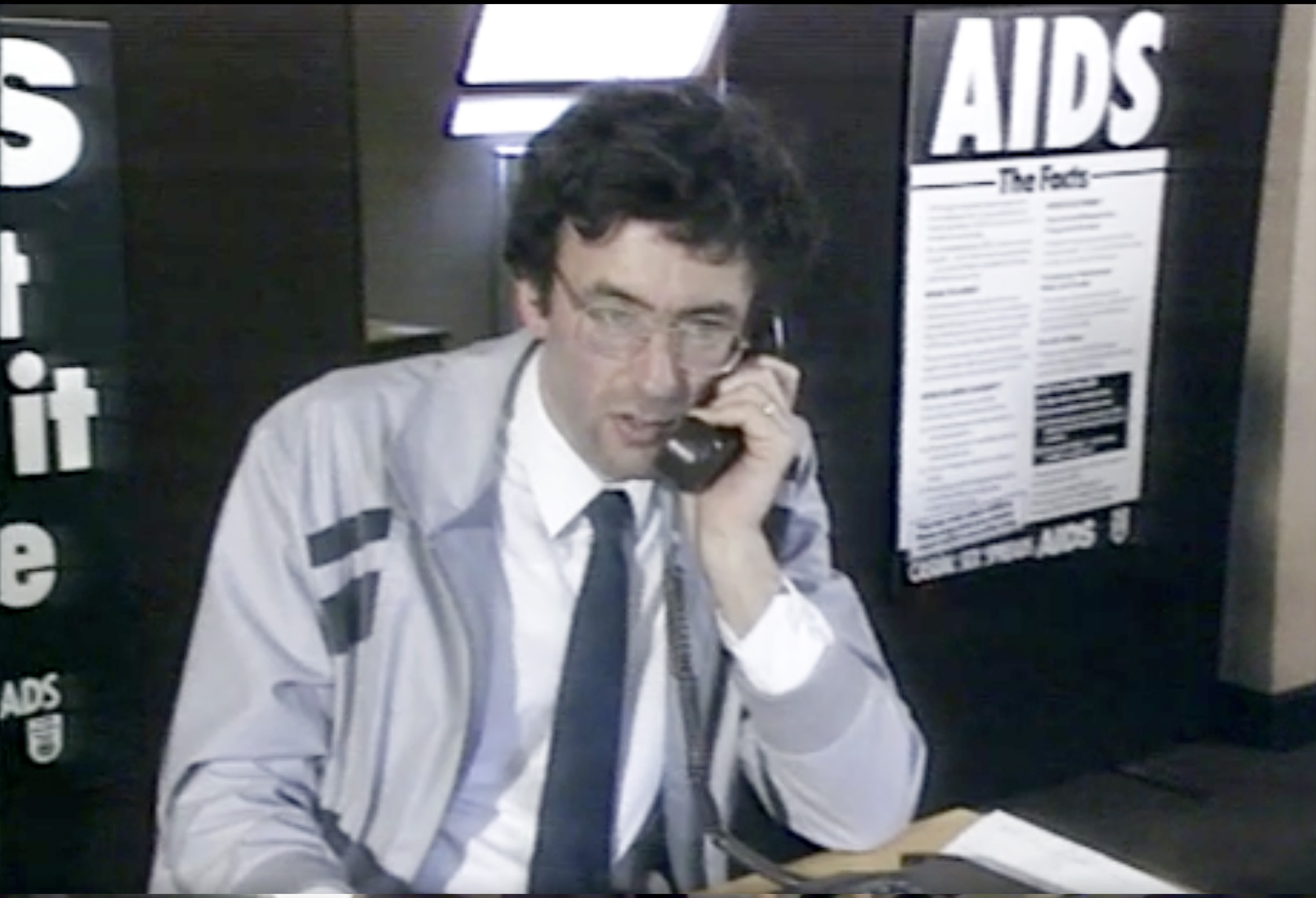A man in a grey suit sits at a desk on the telephone.
