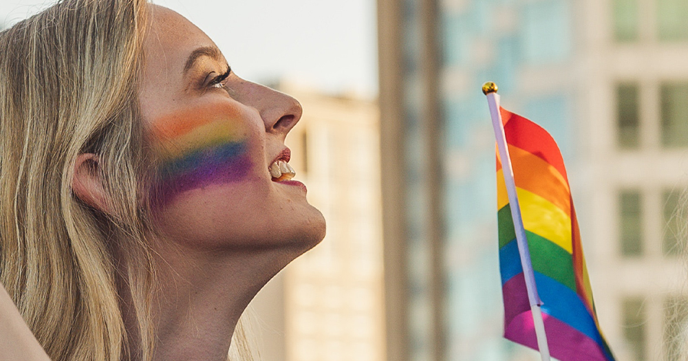 A smiling woman holding a Pride flag with a rainbow painted on her cheek