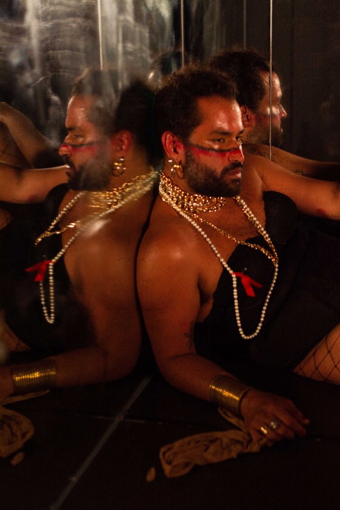 A man slouches against a mirror while wearing a pearl necklace