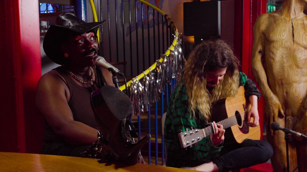 Two performers sit together one in a cowboy hat with a microphone, the other with a guitar