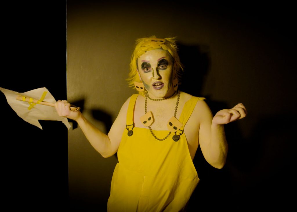 A drag artist stares at the viewer with arms up dressed in yellow