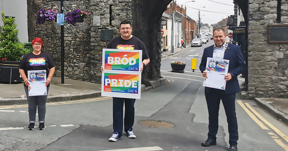3 people stand in front of St. Laurence's Gate to launch Drogheda Pride, holding rainbow postcards and posters