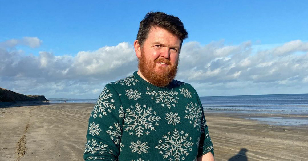 A bearded man wearing a Christmas jumper on a beach - James O'Hagan the author of the Instagram post named 