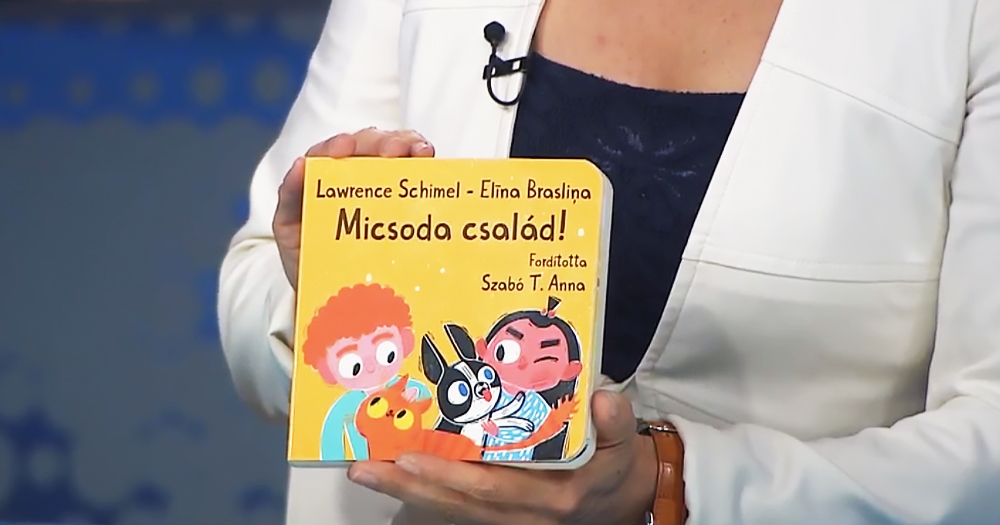 A person holding a copy of a Hungarian LGBTQ+ children's book. The book has a yellow cover and depicts two children as well as two pet animals.
