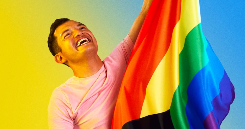 A man laughing while holding a rainbow flag. In this piece, Jorge shares his coming out journey