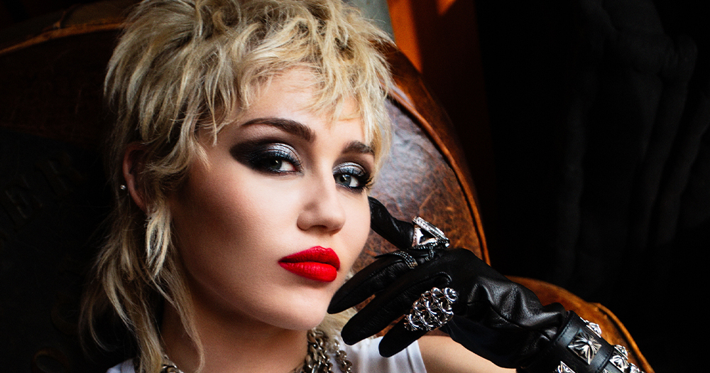 A close-up portrait style photo of Miley Cyrus, with blonde hair, a smokey-eye makeup look, and red lipstick. Cyrus is wearing black leather gloves and she is sitting on a brown leather seat.