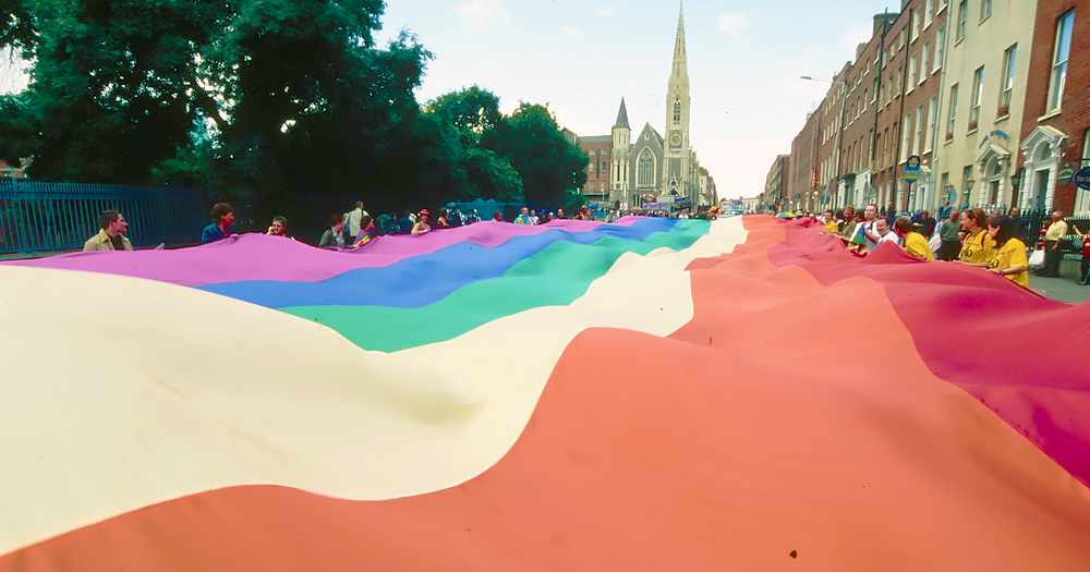 A rainbow LGBTQ+ Pride flag billows over a street. In the background you can see a church and several other buildings.