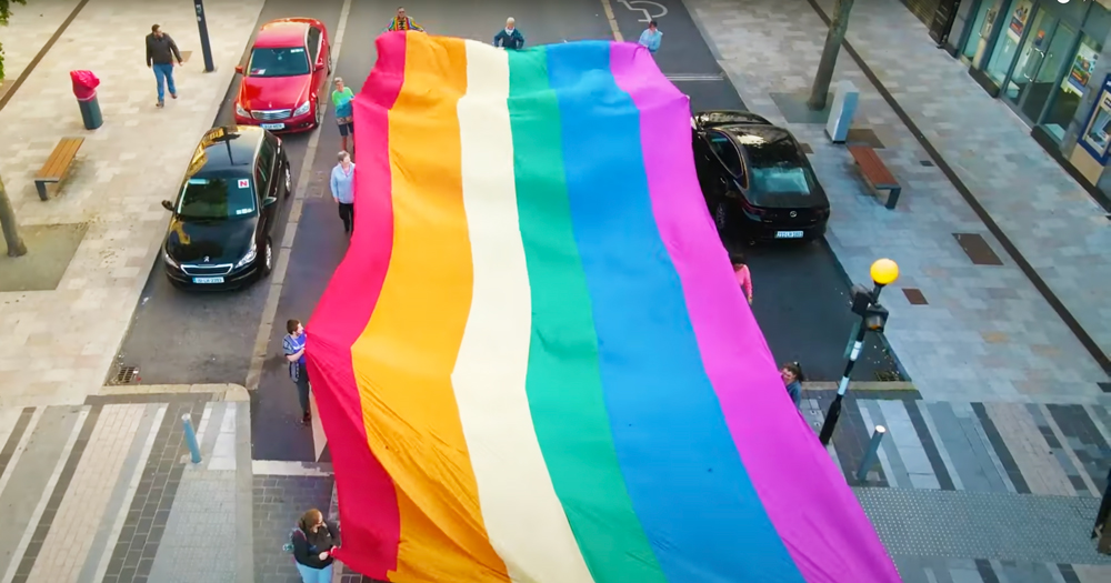a still from 'now you see us' shows a large Pride flag being carried by a group of people down a street