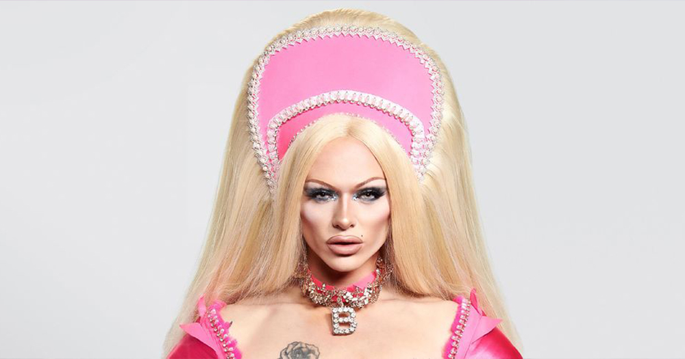 A close up of Drag Queen Bimini, in their promo look for Drag Race UK; a baby pink headpiece, blonde hair and choker with the letter 'B' - Bimini will be guest DJing on BBC Radio 1 for Drag Day