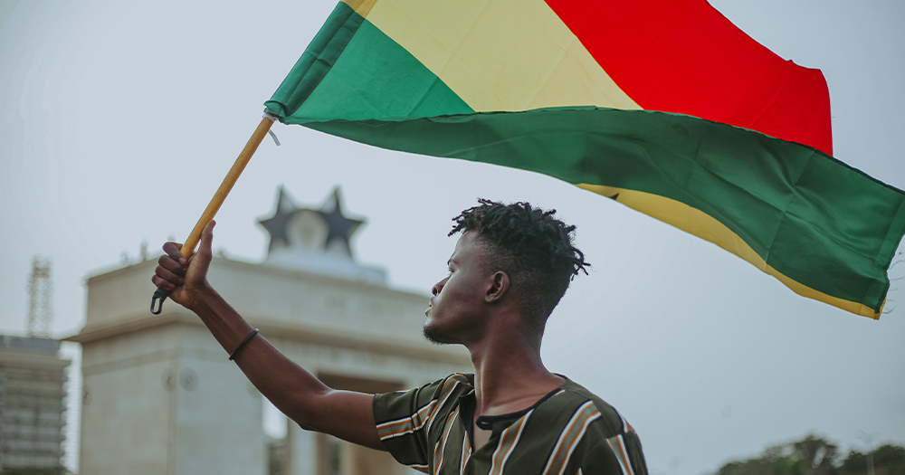 A photo of a man flying the Ghanaian flag. There is a government building in the background.