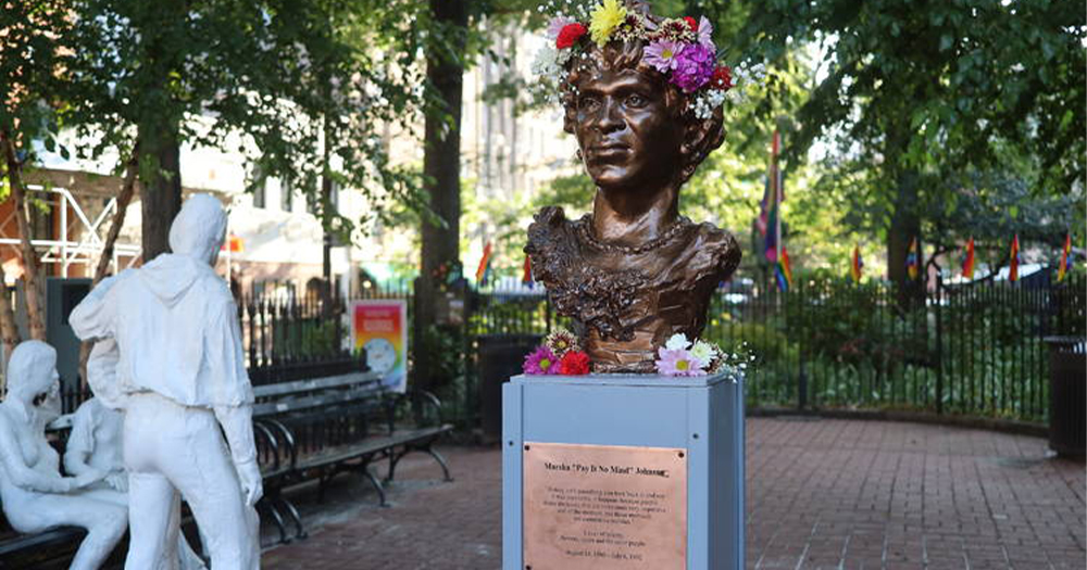 The statue of Marsha P Johnson is shown with flowers in her hair. George Segal’s ‘Gay Liberation’ monument is seen to the left.