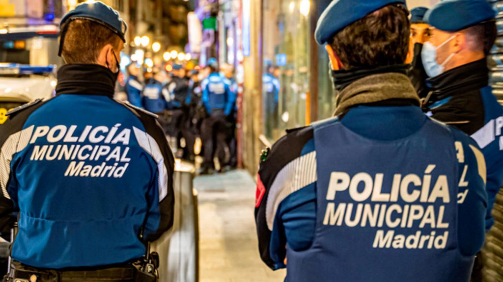 Three Spanish police officers have their backs to the camera. They are standing on a busy street at night time.
