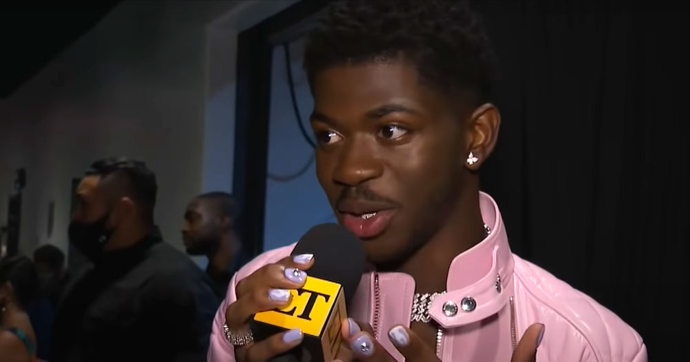 Lil Nas X backstage at awards event, wearing a pink jacket and painted nails, speaking into a microphone, with others in background