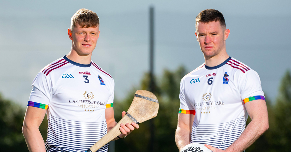 Two young men wear the new GAA Pride Jersey. The strip is white with navy stripes on the lower half and rainbow colours on the cuffs.