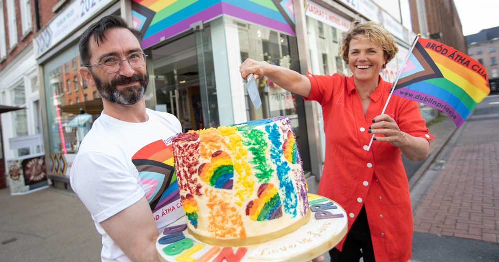 Bearded man holding a colourful cake with women dressed in red holding a knife above the cake while waving a miniature Pride flag. They are standing in front of a shop marked with Pride flag.