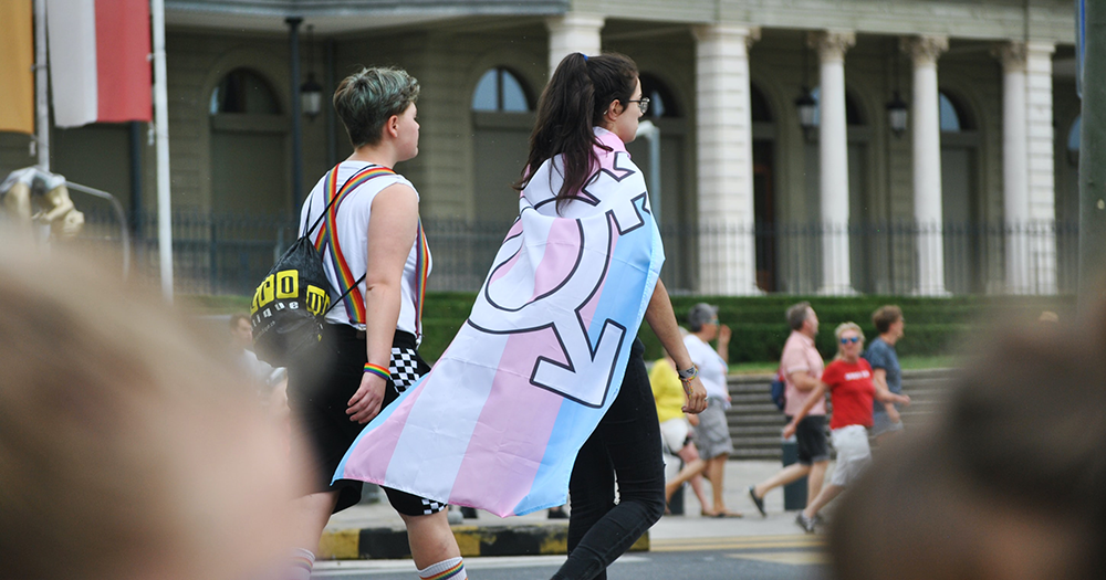 A person wearing the Trans flag around their neck walks proudly through a street as the the LGBTQ+ community comes together to denounce the latest BBC anti-Trans article.