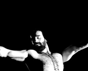 Black and white image of a man from the chest up. He is topless with his arms outstretched and his eyes closed.