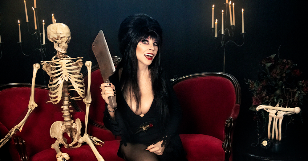 Elvira releases her special with a publicity photo featuring a gothic woman holding a cleaver sitting on a couch beside a skeleton