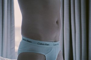 Mid of a man's torso standing in front of a window with grey curtains. He is naked except for white briefs. The waste band reads Calvin Klein.