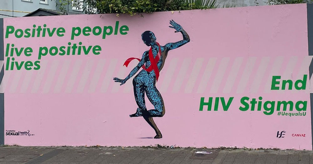 Photograph of the Cork mural for World AIDS Day 2021. The mural is painted onto a light pink background with a dancing grey figure wrapped in a red AIDS ribbon. On the wall is green text which reads 