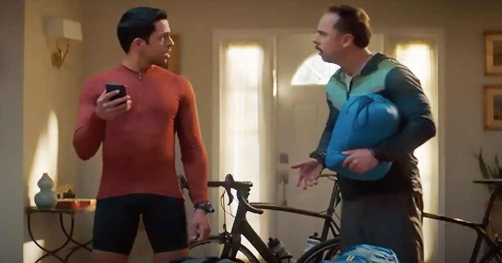 Two men look at each other in discussion behind a bike. One is holding a phone, and the other is holding a bag. These two men are being featured in a queer commercial.